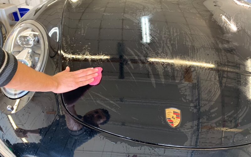 Reinigungsknete wird von Hand über die schwarze Motorhaube eines Porsche geführt